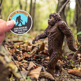 A hand holding a Believe In Bigfoot sticker in front of Bigfoot!