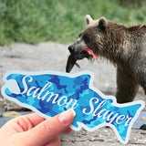 A pic of a bear with a salmon in its mouth and a hand holding a salmon slayer sticker in front of it. 