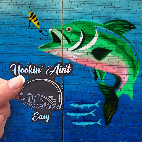 A Hookin' Ain't Easy sticker in front of a brick wall with a mural of a fish on it. 