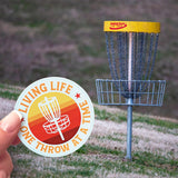 A Living Life One Throw At A Time sticker in front of a disc golf basket. 