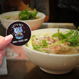 A couple of bowls of pho near a Let's Get Pho-cked Up! sticker. 