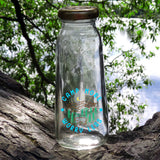 This is a picture of the Camp More Worry Less clear sticker on a tall glass bottle that is sitting on a log outside. 