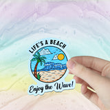 A pic of a hand holding a Life's A Beach sticker against a colorful background!